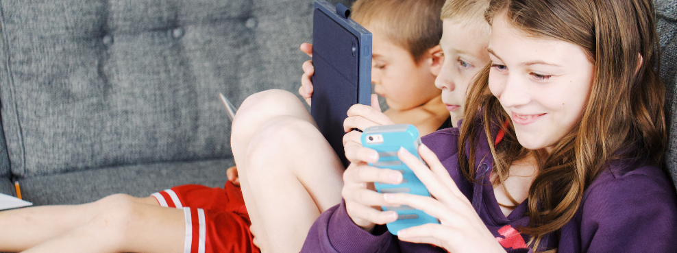 Drei Kinder sitzen mit Handys und Tablets auf dem Sofa.