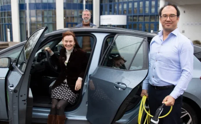 Eine Frau sitzt in einem E-Auto und schaut durch die geöffnete Tür, ein Mann steht vor dem Auto, einer dahinter.