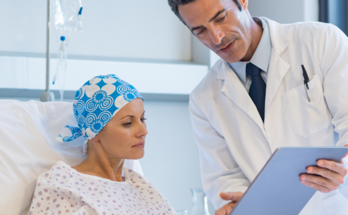KI-gestützte Wearables sollen helfen, Komplikationen bei der Therapie von Krebspatient:innen schneller zu erkennen. (Foto: Canva) Ein Arzt hält einer Krebspatientin ein Klemmbrett hin und erklärt ihr etwas.