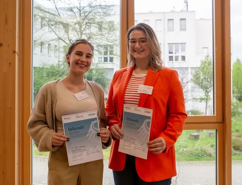 Studierendenförderung mit dem Deutschlandstipendium (Foto: UW/H │ Lucy Midnich) Foto von zwei Frauen, die nebeneinander vor einem Fenster stehen. Beide halten eine Urkunde in den Händen.