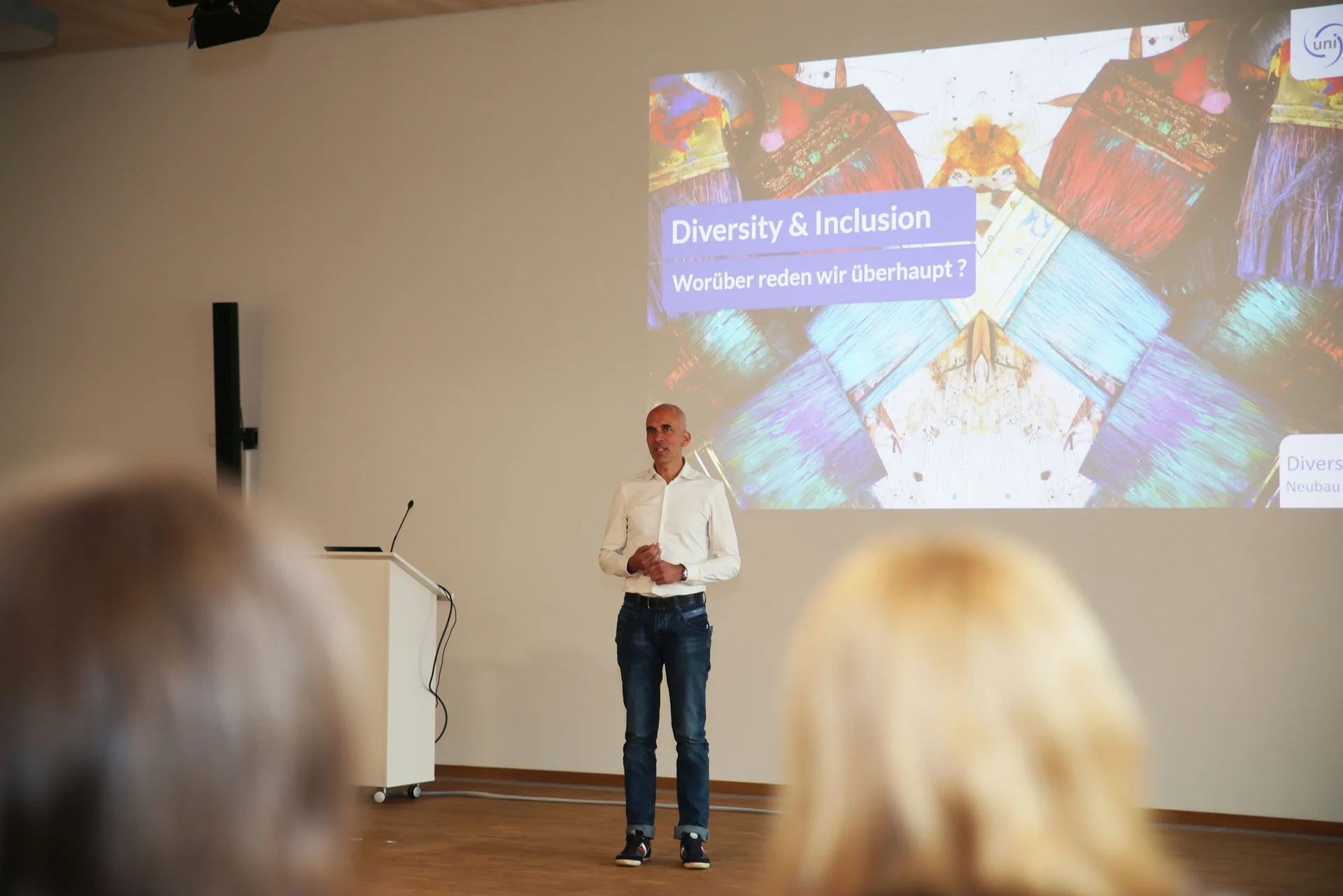 Dr. Dirk Jakobs, Vizepräsident für Organisationsentwicklung an der UW/H, steht auf der Bühne neben einem Rednerpult und spricht zum Publikum. Das Veranstaltungstitelbild ist als Projektion an der Wand hinter ihm zu sehen. 