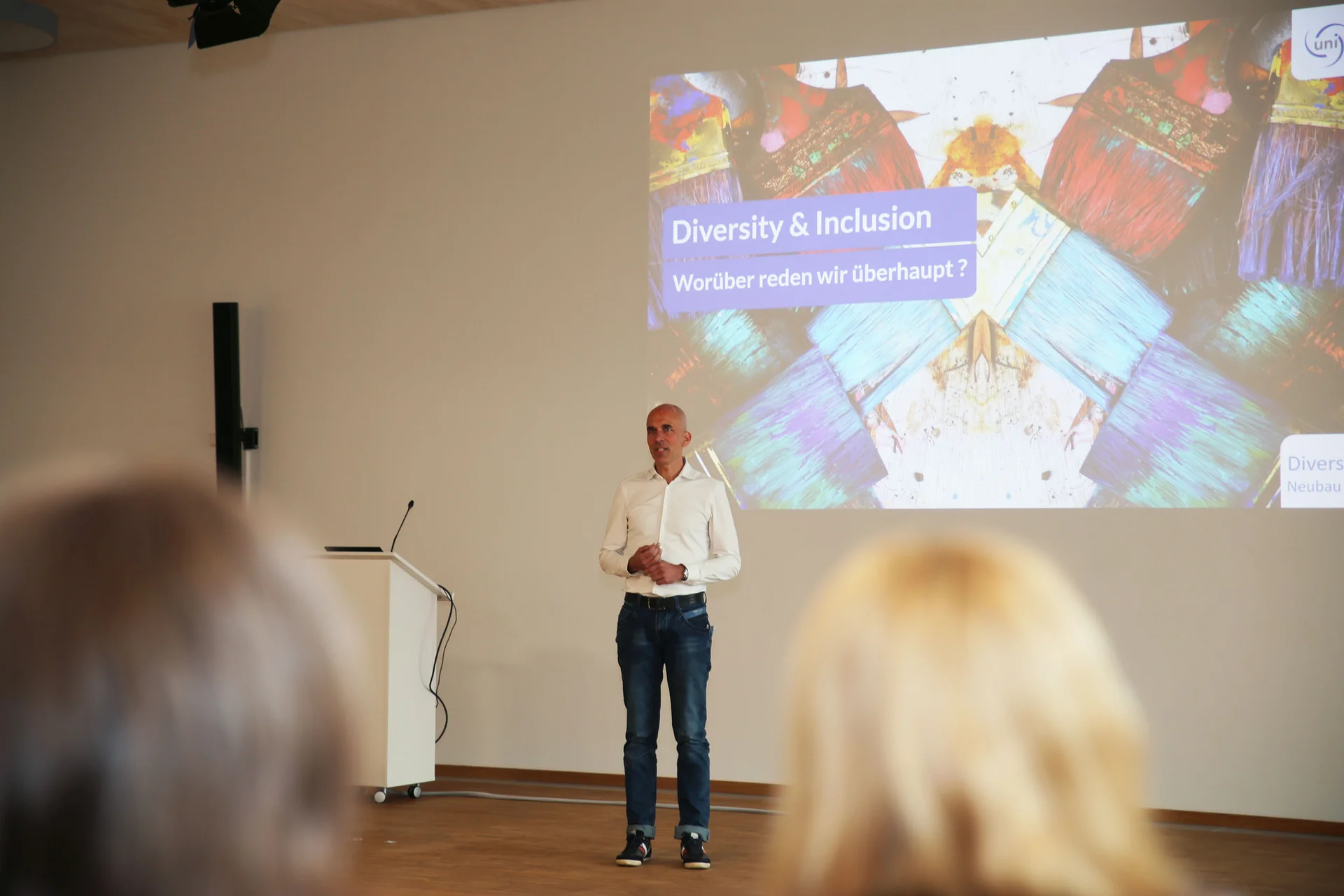 Austausch beim Diversity-Tag an der UW/H (Foto: UW/H) Dr. Dirk Jakobs, Vizepräsident für Organisationsentwicklung an der UW/H, steht auf der Bühne neben einem Rednerpult und spricht zum Publikum. Das Veranstaltungstitelbild ist als Projektion an der Wand hinter ihm zu sehen.