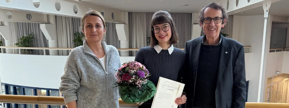 Three people stand next to each other in the university. In the centre is Theresa Busse, a young woman holding her appointment certificate and a bouquet of flowers and looking happy. To her left is Prof Dr Margareta Halek, to her right Prof Dr Stefan Zimmermann.
