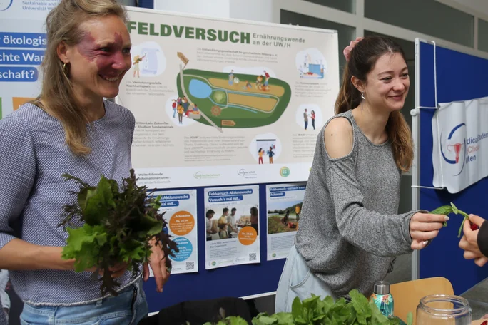 Das Projektteam stellt sich vor (Foto: UW/H) Zwei Frauen präsentieren ihr Projekt. Eine von ihnen hält einen Kopfsalat in den Händen. Hinter ihnen ist eine Pinnwand mit Infoplakaten.