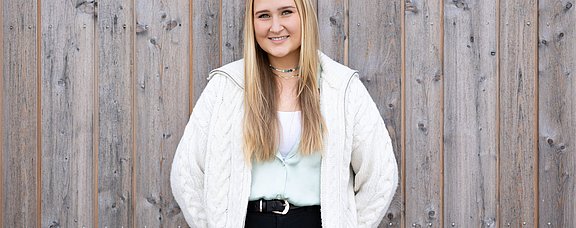 Bewirb dich für den Studiengang Psychologie (M. Sc.) an der UW/H. (Foto: UW/H | Michael Schwettmann) Eine junge Frau steht mit dem Rücken vor einer Gebäudewand aus Holz.