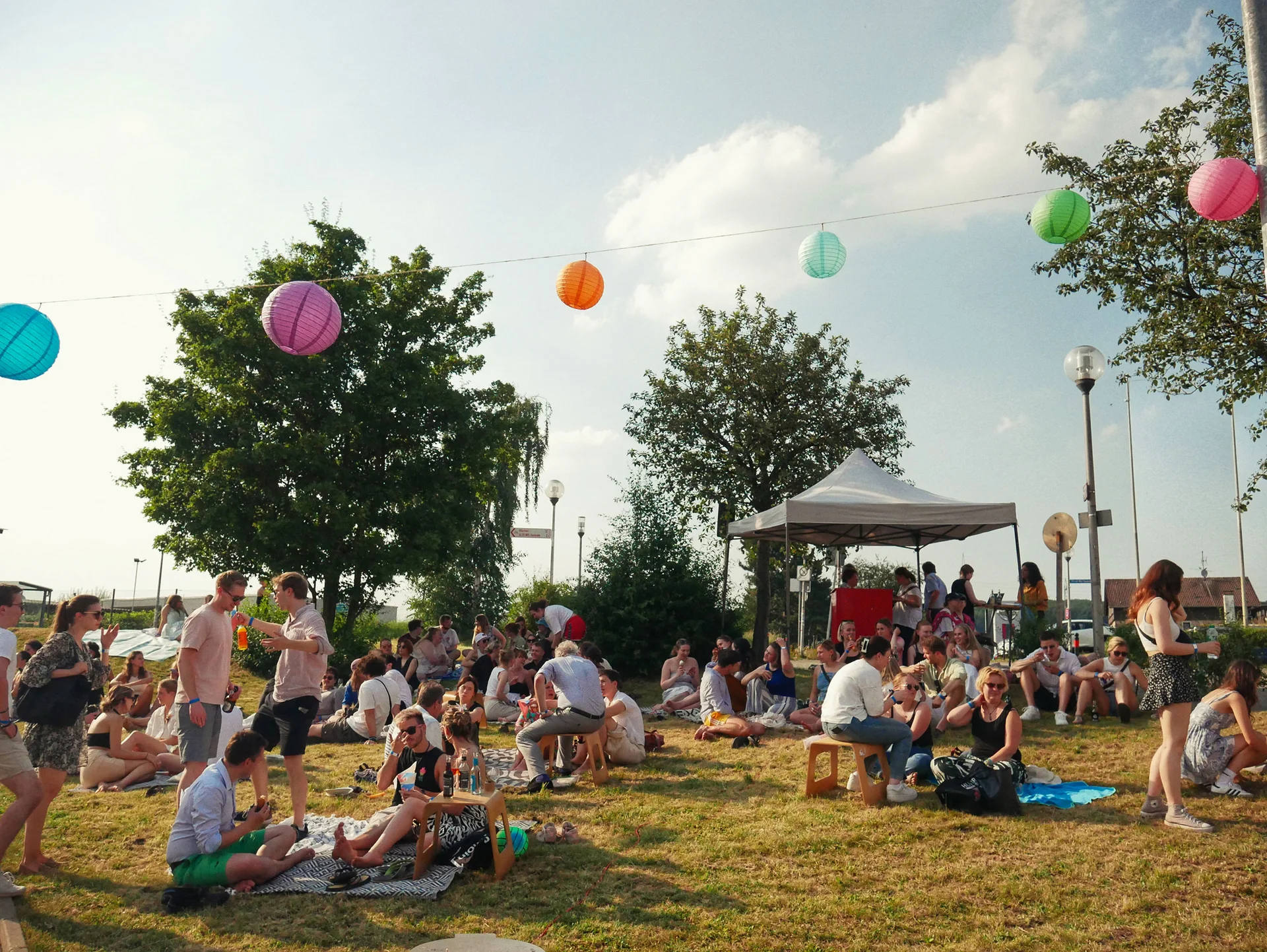 Celebrating together at the summer party (Photo: UW/H | Cara Wegerer). People are sitting or standing on a meadow. A garland of colourful lanterns is strung above them.