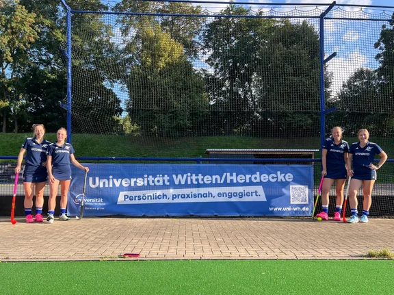 Vier Hockey-Spielerinnen flankieren ein blaues Werbebanner der Uni Witten/Herdecke am Spielfeldrand eines Hockeyfeldes.
