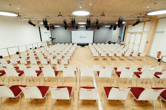 Großer Veranstaltungsraum mit Platz für bis zu 350 Zuschauer:innen (Foto: UW/H | Michael Kotowski) Aufnahme eines Saals, in dem rotgepolsterte Stühle mit weißen Lehnen in Reihen stehen, vorne ist eine Leinwand angebracht