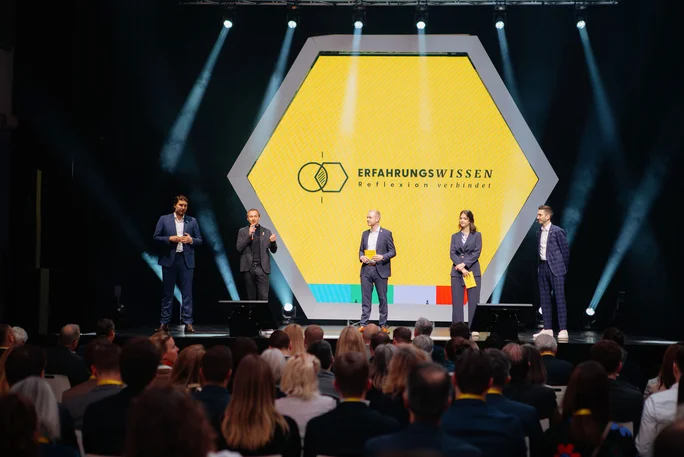 Zu sehen ist ein Bühnenvortrag mit fünf Personen bei einer Veranstaltung