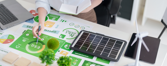 Ansicht eines Schreibtisches, auf dem verschiedene Unterlagen mit Diagrammen und Statistiken liegen und außerdem Miniatur-Bäume und -Windräder stehen.