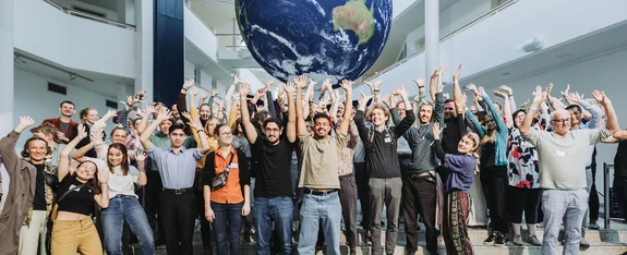 A large group of people stand under an inflatable globe and raise their arms in the air.