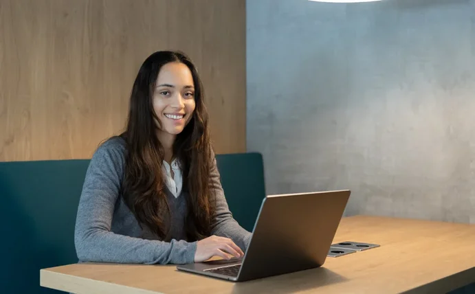 Eine Studentin sitzt mit ihrem Laptop an einem Tisch und lächelt in die Kamera.