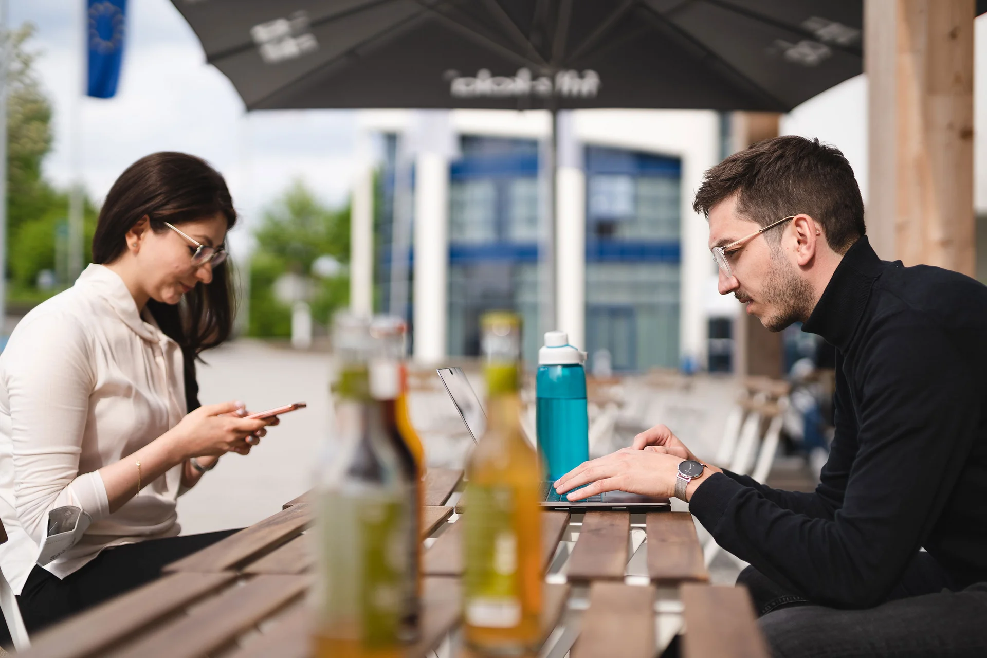 Two students sit opposite each other at a wooden table. They are looking at their mobile phones or laptops. Drinks are in front of them. The university building is in the background.