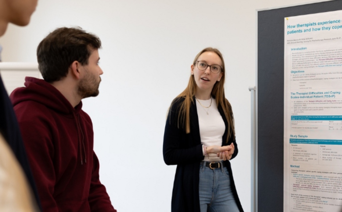 Studierende stehen vor einer Pinnwand, auf der Psychologische Modelle beschrieben sind. Es handelt sich um eine Lerngruppe.