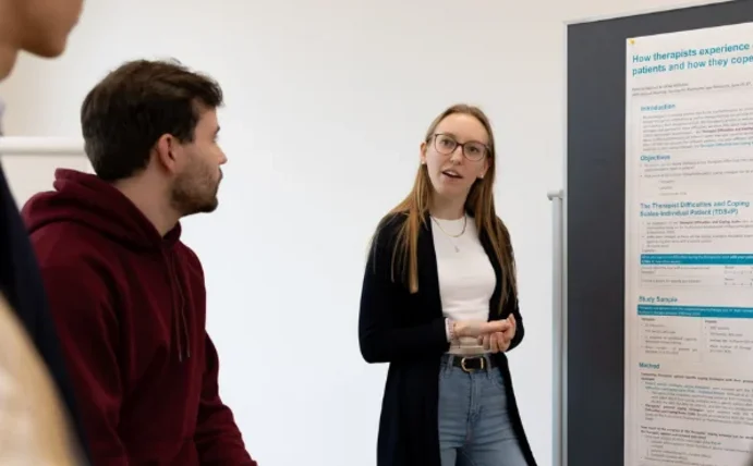 Studierende stehen vor einer Pinnwand, auf der Psychologische Modelle beschrieben sind. Es handelt sich um eine Lerngruppe.