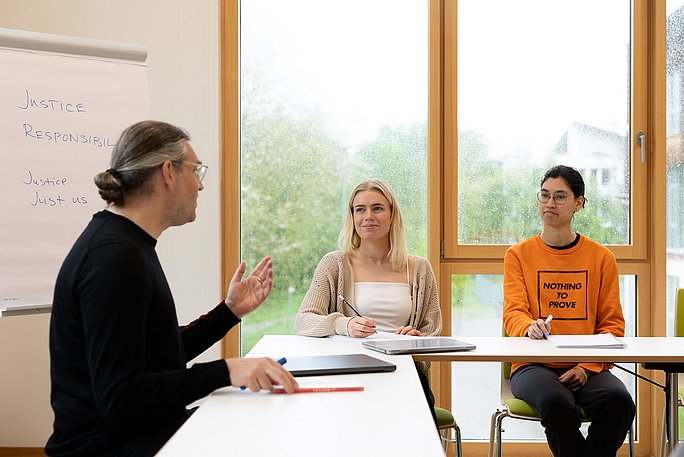 Ein Professor sitzt mit zwei Studierenden im Seminarraum und diskutiert.