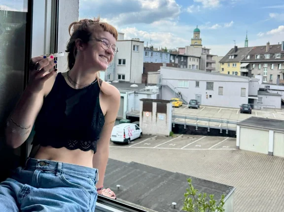 Foto von Emmi Mentzel am Fenster ihrer WG. Im Hintergrund sind ein Hinterhof und mehrere Gebäude zu sehen.