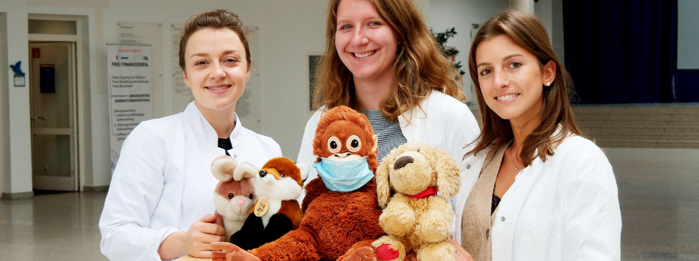 Drei Studentinnen lächeln in die Kamera. Auf einem Tisch vor ihnen befinden sich mehrere Stofftiere.