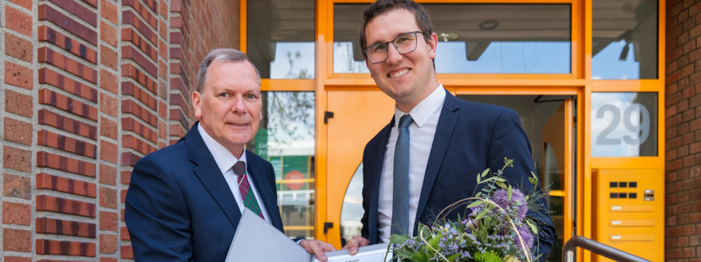 Dr. Fabian Zurnieden wurde für seine Forschung zur zahnmedizinischen Versorgung von Menschen mit Behinderung in betreuten Wohnformen ausgezeichnet. (Foto: OralGesundheit Inklusiv – OGI) Zwei Männer lächeln in die Kamera und halten gemeinsam eine Urkunde hoch. Der Mann rechts hat außerdem einen Blumenstrauß in der Hand.
