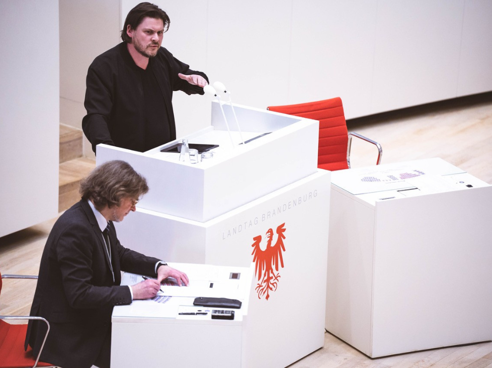 Oliver Skopec steht hinter einem weißen Rednerpult, auf dem das Brandenburger Wappen abgebildet ist.