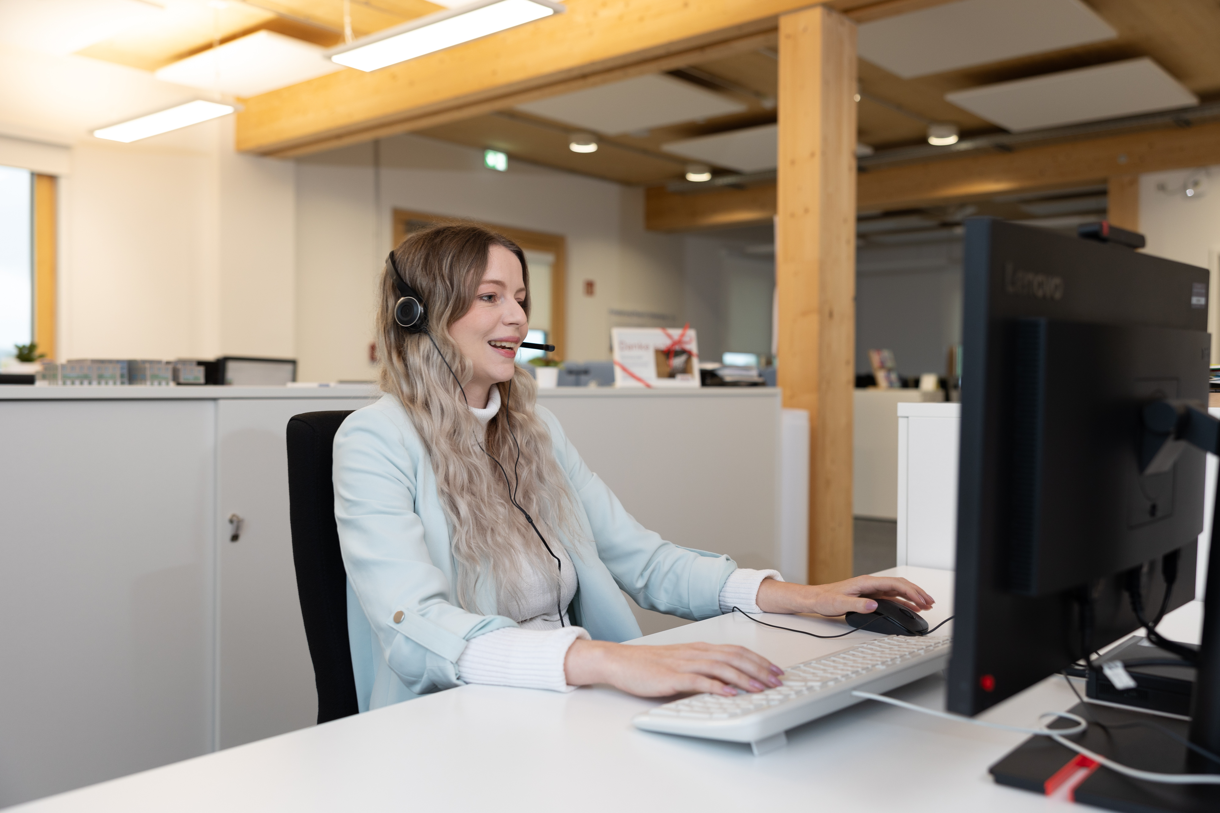 Eine junge Frau mit einem Headset sitzt in einem Großraumbüro vor einen Computer.